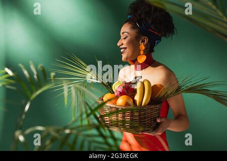 Lächelnde junge afroamerikanische Frau in rotem Badeanzug mit Korb mit exotischen Früchten auf Grün Stockfoto