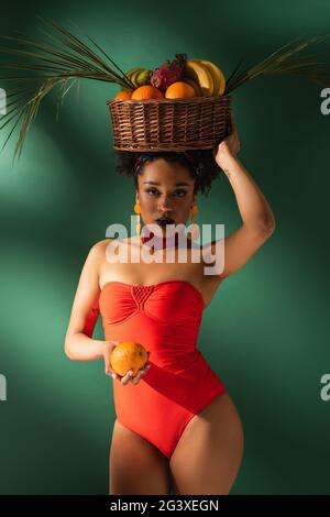 Junge afroamerikanische Frau in rotem Badeanzug mit Korb mit exotischen Früchten auf Grün Stockfoto