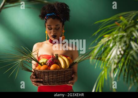 Junge afroamerikanische Frau, die Korb mit exotischen Früchten auf Grün betrachtet Stockfoto