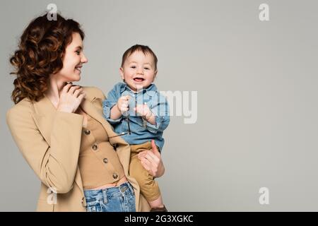 Fröhlicher Kleinkind Junge hält Brille, während glückliche Mutter in Blazer lächeln isoliert auf grau Stockfoto