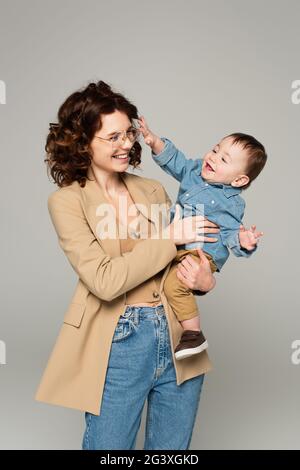 Fröhliche Mutter in der Brille und Blazer hält in den Armen glücklich Baby Junge isoliert auf grau Stockfoto