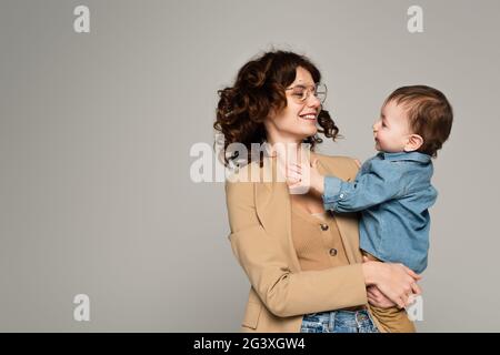 Glückliche Mutter in Brille und Blazer hält in den Armen Baby Junge isoliert auf grau Stockfoto