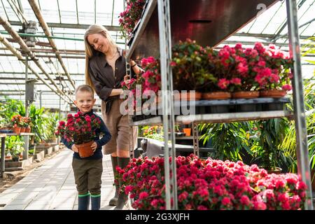 Kleiner Junge mit einer Blume in einem Topf und seine Mutter, eine Floristin, die einen Karren mit Blumen in einem Gewächshaus zieht Stockfoto