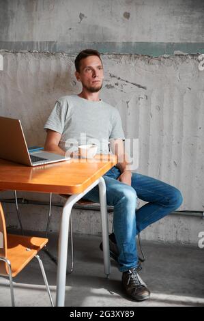 Mann sitzt am Tisch mit Laptop und einer Tasse Kaffee. Junger Mann ruht sich nach der Arbeit online aus. Konzept des Heimbüros. Stockfoto