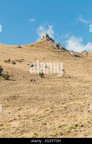 Verlassene Herbstebene in Bergen mit vergilbtem Gras. Felsen an einem Hang in Bergen. Bergige Gegend vor blauem Himmel. Stockfoto