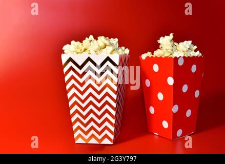 Popcorn in farbiger Kartonverpackung. Haufen gesalzenes Popcorn in Papier gestreiften Eimer auf rotem Hintergrund. Stockfoto