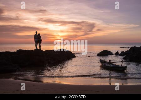 Silhouette eines jungen Paares auf Felsen und Fischerboot am Ong lang Beach bei Sonnenuntergang, Ong lang, Phu Quoc Island, Kien Giang, Vietnam, Asien Stockfoto