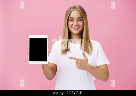 Eine Aufnahme einer optimistischen und fröhlichen Frau in der Taille, die ein cooles digitales Tablet zeigt, das auf den Bildschirm des Geräts zeigt und breit auf den Kameralinder lächelt Stockfoto