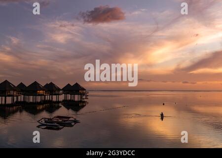 Silhouette einer Frau im Wasser neben dem Auslegerkanu und den Überwasser-Bungalows des Tahiti Ia Ora Beach Resort (verwaltet von Sofitel) bei Sonnenuntergang, Stockfoto