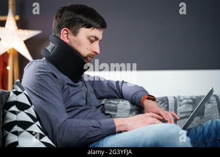 Ein Mitarbeiter, der nach einem Unfall zu Hause hinter einem Laptop arbeitet und Schmerzen im Nacken spürt, verwendet eine schwarze Nackenstütze. Männlich mit Hals Stockfoto
