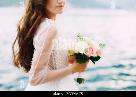 Die Braut hält einen Strauß Rosen in der Hand und steht auf dem Pier in der Bucht von Kotor, aus nächster Nähe Stockfoto