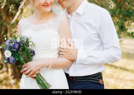 Der Bräutigam umarmt die Braut sanft in der Nähe der grünen Bäume im Olivenhain, die Braut hält einen Strauß blauer Blumen Stockfoto