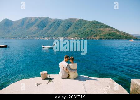 Ein Mann und eine Frau sitzen und umarmen am Rande eines Piers in der Nähe von Perast in der Bucht von Kotor, Rückansicht Stockfoto