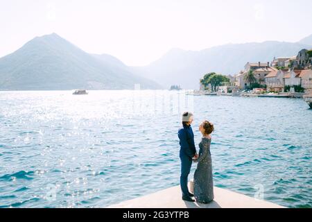 Die Braut in einem stilvollen grauen Hochzeitskleid und die Der Bräutigam steht mit den Händen an der Küste in der Nähe des alten Stadt Perast Stockfoto