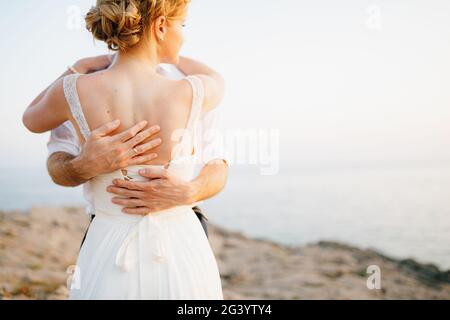 Die Braut und der Bräutigam umarmen sich auf den Felsen am Meer vor der Kulisse der Berge und der Insel Mamula, Rückansicht Stockfoto