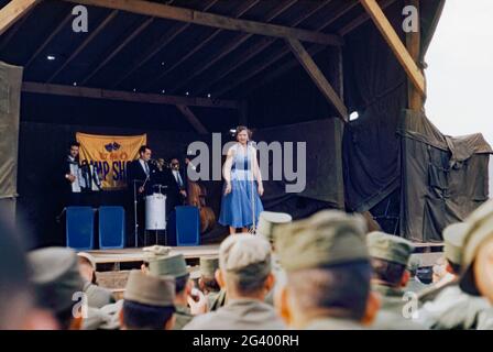 Eine ‘„USO Camp Show“, die amerikanische Truppen während des Koreakriegs unterhielt, Südkorea, 1954. Die Sängerin wird von einer dreiköpfigen Band unterstützt und das USO-Banner steht auf der temporären Bühne hinter ihnen. 1954 besuchte Marilyn Monroe Korea, um die Truppen zu unterhalten. Auch in diesem Jahr tourte die ‘Slapsie Maxie’ Show. The United Service Organizations Inc (USO) ist eine amerikanische Wohltätigkeitsorganisation, die den US-Streitkräften Live-Unterhaltung wie Komiker, Schauspieler und Musiker bietet. Dieses Bild stammt von einer Kodak-Amateurfarbtransparenz, die von einem Mitglied des Publikums aufgenommen wurde. Stockfoto