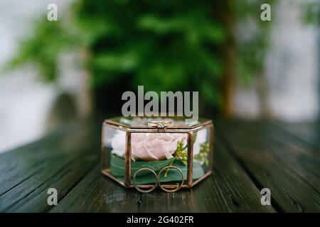 Der Verlobungsring und die Eheringe der Braut und des Bräutigams in einer dekorativen Glasbox mit einer rosa Blume im Inneren auf einem verschwommenen BA Stockfoto