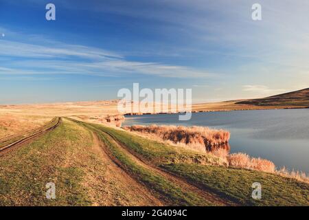 Großer blauer See, der im Herbst mit Schilf bewachsen ist. Ländliche Landschaft mit einer Landstraße gegen den blauen Himmel. Gelbes Schilf in der Au Stockfoto