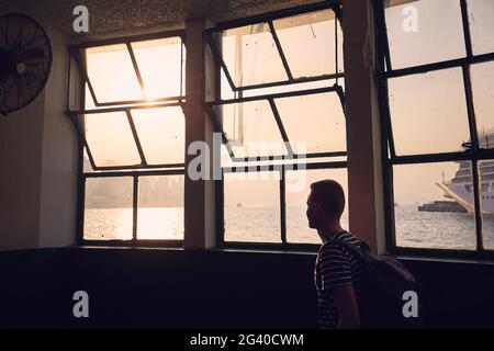 Nachdenklicher Mann, der beim Sonnenuntergang durch das Fenster des Fährenterminals auf die Skyline der Stadt blickt. Silhouette des Touristen in Hongkong. Stockfoto