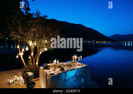Romantisches Abendessen für zwei Personen an einem malerischen Ort mit einem Tisch mit Kerzen und Tellern zum Essen mit Blick auf das Wasser und die Berge Stockfoto