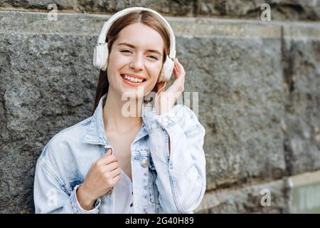 Ein junges Mädchen mit Kopfhörern hört Musik und posiert freudig gegen eine steinerne, graue Wand Stockfoto