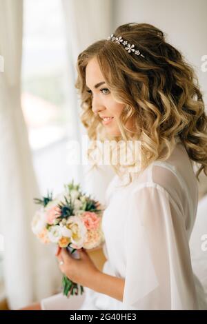 Eine Braut in einem sanften Peignoir mit einem Blumenstrauß in den Händen sitzt am Fenster mit weißen Vorhängen und lächelt, Nahaufnahme Stockfoto