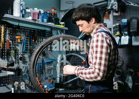 Männlicher Mechaniker, der in der Fahrradwerkstatt arbeitet, Mechaniker, der Fahrrad mit einem Spezialwerkzeug repariert und Schutzhandschuhe trägt. Junge Attra Stockfoto