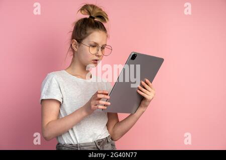 Nettes blondes Mädchen mit Brille, die auf das Tablet schaut. Teen isoliert auf einem rosa Hintergrund. Stockfoto