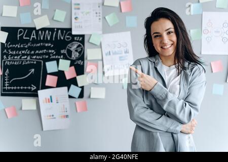 Lächelndes junges Mädchen im Büro auf einem Hintergrund einer grauen Wand mit Aufklebern. Sie zeigt mit ihrem Zeigefinger auf etwas. Stockfoto