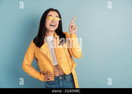 Das positive, lächelnde Mädchen zeigt mit dem Zeigefinger nach oben auf den Kopierbereich. Schöne, junge Frau in Sonnenbrillen isoliert auf blauem Hintergrund. Stockfoto