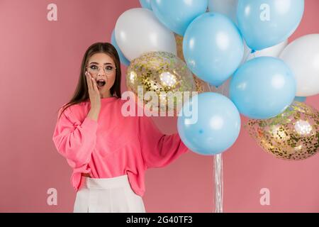 Schockiert, überrascht Mädchen in Brille hält eine Menge Ballons, berührt ihr Gesicht. Mädchen in einer rosa Jacke auf einem rosa Hintergrund, schockiert, überrascht Mädchen in g Stockfoto