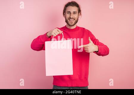 Ein hübscher, bärtiger Mann zeigt auf eine Papiertüte. Lächelnder Kerl auf einem rosa Hintergrund zeigt seinen Finger auf einer leeren Papiertüte, Platz für Text, Kopierraum. Stockfoto