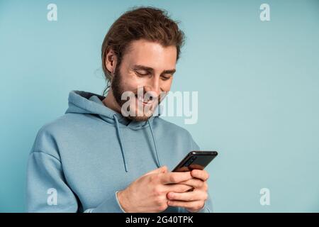 Lächelnder Mann mit Bart, in einem bequemen Kapuzenpullover, mit einem Telefon in den Händen. Mann auf einem rosa Hintergrund. Stockfoto