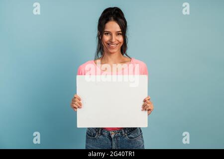 Attraktive Brünette hält in ihren Händen ein leeres, zerschlagene Blatt. Mädchen in einem rosa T-Shirt auf blauem Hintergrund hält sie ein weißes Stück Papier zum Kopieren Stockfoto