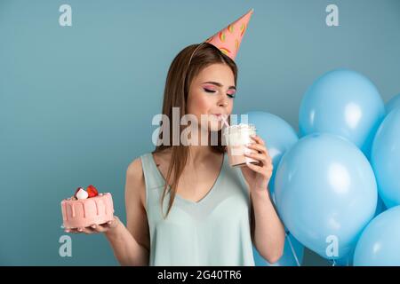 Schönes, elegantes Geburtstagsmädchen in einem festlichen Hut singt ihren Cocktail und hält einen Weihnachtskuchen. Mädchen posieren vor dem Hintergrund von Luftballons und einem blu Stockfoto