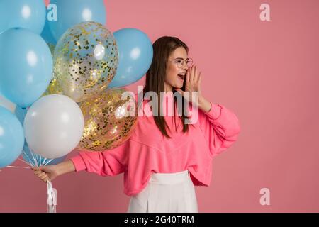 Nette, schöne, junge Frau, die viele Ballons in der Hand hielt, ihre Hand ins Gesicht legte, etwas lautes sagte. Schönes Mädchen posiert auf einem rosa Hintergrund, Stockfoto