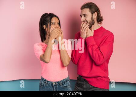 Das junge Paar eines Jungen und eines Mädchens schloß mit den Händen den Mund und schaute einander an. Nettes Mädchen und Kerl auf einem rosa und blauen Hintergrund Stockfoto