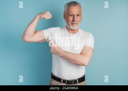 Älterer bärtiger Mann, der Muskeln in weißer T-Shirt-Pose auf blauem Studiohintergrund zeigt. Ernsthafter Mann weist auf seinen perfekten Bizeps hin Stockfoto