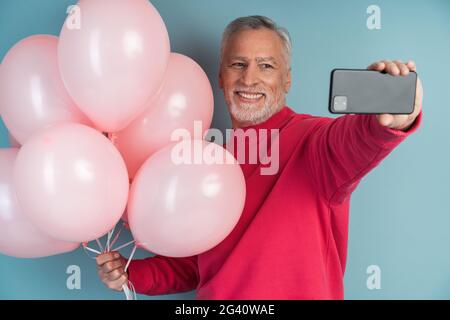 Fröhlicher, lächelnder, grauhaariger Mann mit Kugeln auf blauem Hintergrund. Positive Person, die ein Selfie am Telefon macht, fröhlich, lächelnd, grauhaariger Mann w Stockfoto