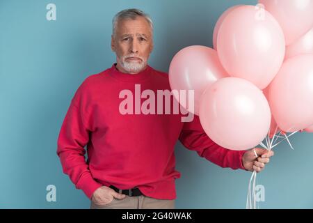 Verärgert, älterer bärtiger Mann trägt, Ballone in der Hand haltend. Älterer Mann, der auf blauem Hintergrund posiert, verärgert, älterer bärtiger Mann trägt und Luftballons in der hand hält Stockfoto