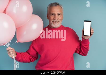 Mann mit Luftballons, die einen hellen Pullover tragen und auf blauem Hintergrund posieren. Netter Mann, der ein Telefon hält, einen leeren, schwarzen Telefonbildschirm zeigt, Platz zum Kopieren, Platz Stockfoto