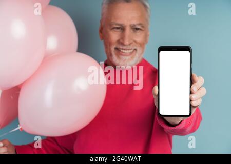 Der süße, lächelnde Großvater hält ein Telefon und zeigt einen leeren, dunklen Bildschirm. Mann hält Ballons auf blauem Hintergrund., netter, lächelnder Großvater hält ein Telefon Stockfoto