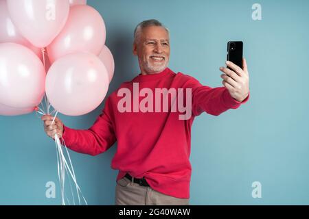 Attraktiver älterer Mann hält Ballons und nimmt ein Selfie auf einem Handy. Mann isoliert auf blauem Hintergrund, Kopierraum, Platz für Text., attraktive seni Stockfoto