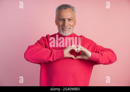 Der ältere Mann zeigt eine Geste aus dem Herzen. Mann liebevoll, ein Herz mit Fingern auf einem rosa Hintergrund, Ort für Text, kopieren Raum Stockfoto