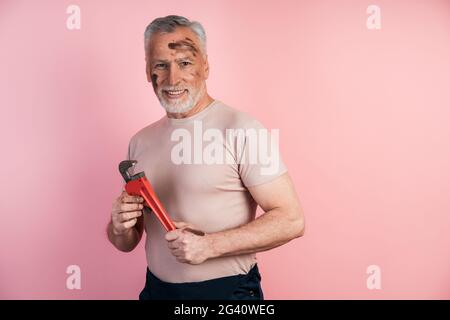 Grauhaariger männlicher Baumeister lächelt in seiner Hand und hält einen Schraubenschlüssel auf einem isolierten rosa Hintergrund. Senior man geölt, Arbeitskonzept., grauhaariger männlicher Buril Stockfoto