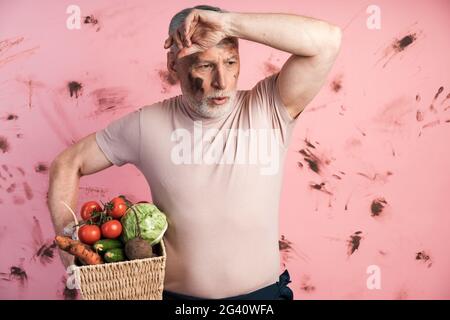 Müde Senior Mann hält einen Korb mit Gemüse auf einem schmutzigen rosa Hintergrund. Ein Mann mit einem verirrten Gesicht nach harter Arbeit., müde älterer Mann mit einem Bas Stockfoto