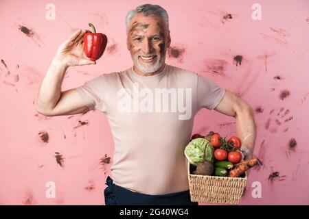 Lächelnd hält ein älterer Mann einen Korb mit Gemüse und einen roten Pfeffer in der anderen Hand. Bauer auf einem schmutzigen rosa Hintergrund., lächelnd, älterer Mann h Stockfoto