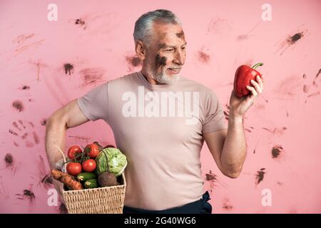 Attraktiver, älterer Mann hält einen Korb mit Gemüse und hält einen roten Pfeffer in der anderen Hand, er untersucht ihn. Bauer auf einem schmutzigen rosa Hintergrund. Stockfoto