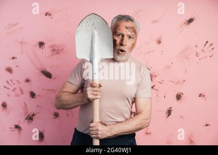 Überrascht blickt der ältere Mann hinter dem Wrack auf einem schmutzigen, rosa Hintergrund heraus. Älterer Mann auf Kopierplatz isoliert. Stockfoto