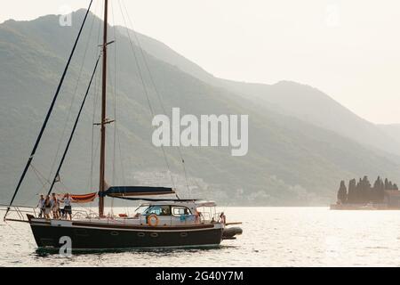 Eine Yacht mit gesenkten Segeln und Menschen an Bord vor der Kulisse der Berge der Stadt Perast in Montenegro. Stockfoto